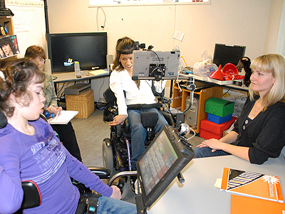 Two students in powered wheelchairs and two adults are seated in a circle inside of a classroom