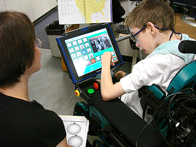 A child in a powered wheelchair uses their AAC device to while their teacher seated behind them looks on 