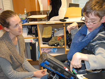 A child in a powered wheelchair using their AAC device to communicate with their teacher sitting in front of them 