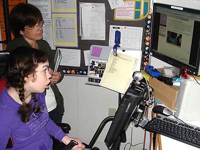 A student in a powered wheelchair and an adult are both looking at a monitor in front of them